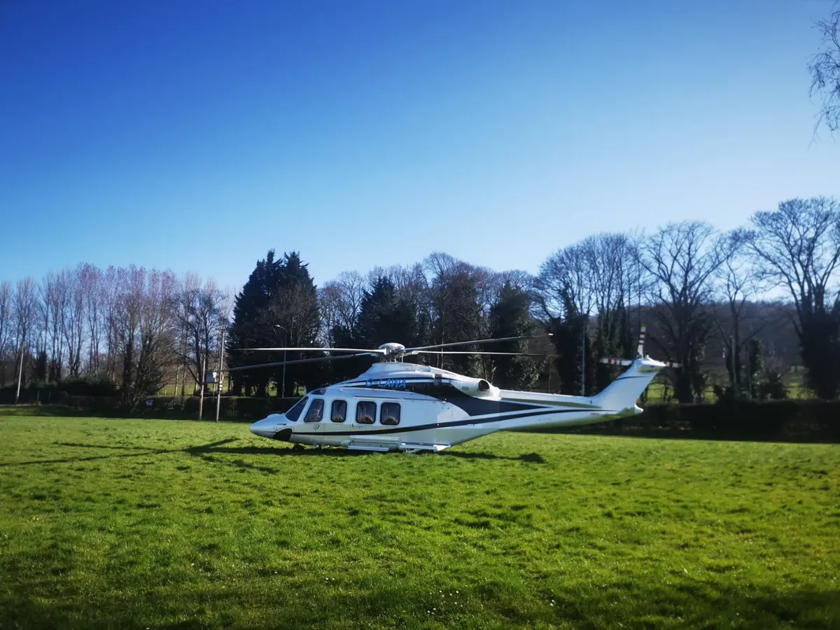 A large white twin-engine helicopter on a grass field among trees